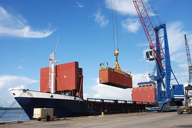A shipping container being loaded onto a cargo vessel at port